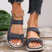 Sandali estivi blu con cinturini in velcro, indossati su piedi con smalto bianco. Jeans strappati e cavigliera dorata. Moda casual donna.
