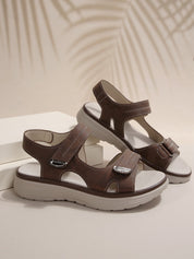 Sandali marroni da donna con cinturini regolabili, suola in gomma antiscivolo, design casual estivo, sfondo neutro con ombre di foglie.