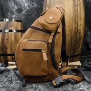 Zaino a tracolla in pelle marrone con tasche multiple, design elegante e robusto, posizionato davanti a barili di legno. Accessorio moda uomo.