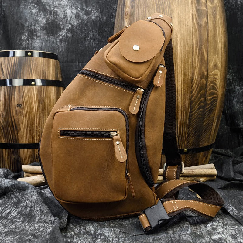 Zaino a tracolla in pelle marrone con tasche multiple, design elegante e robusto, posizionato davanti a barili di legno. Accessorio moda uomo.