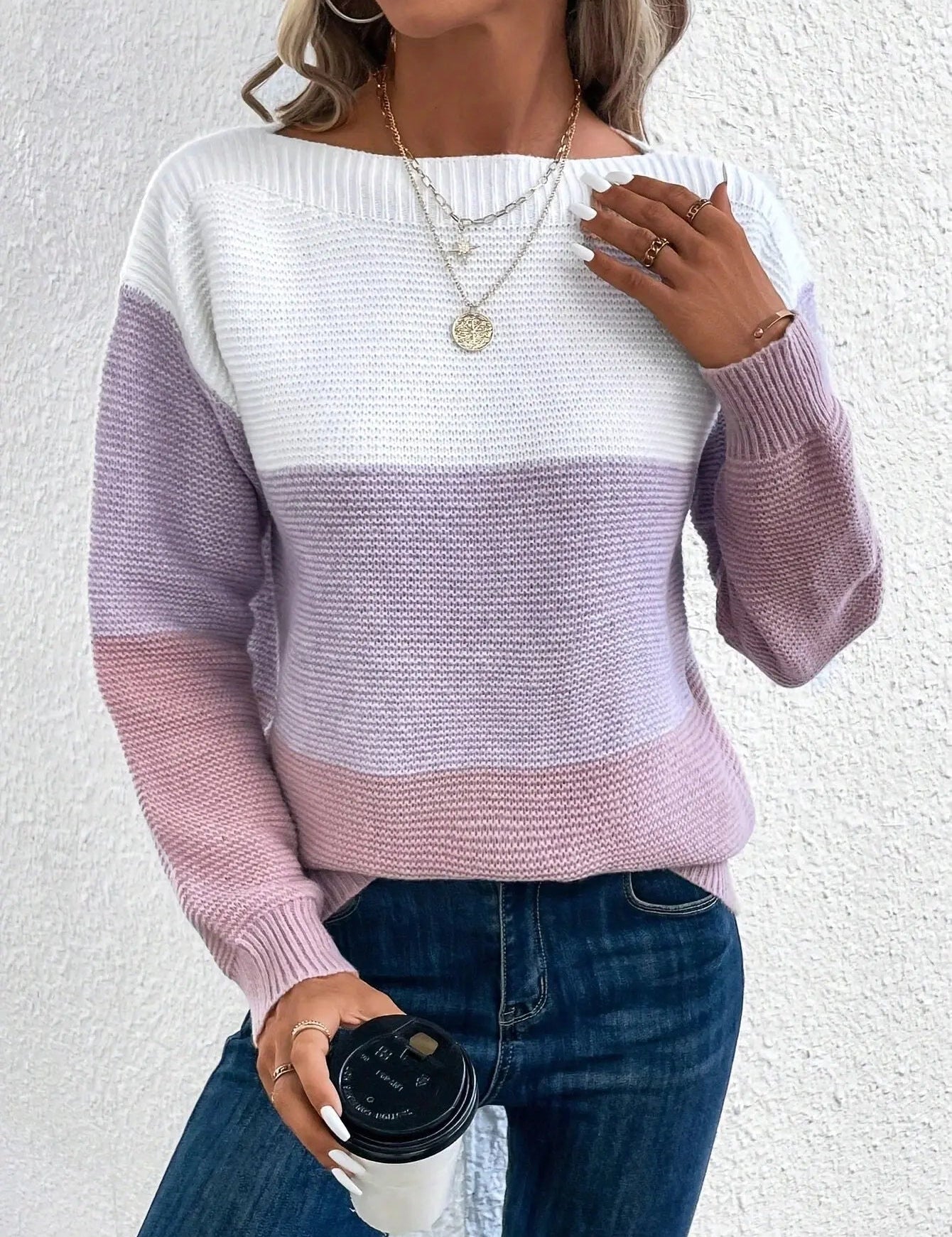 Maglione donna a righe bianche, lilla e rosa, abbinato a jeans blu. Accessori: collane dorate e anelli. Tazza da caffè in mano.