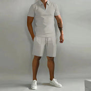 Uomo in completo estivo casual grigio chiaro, polo con zip e pantaloncini, scarpe bianche, tatuaggio sul braccio destro. Moda maschile estiva.
