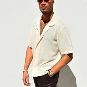 Uomo con camicia a maniche corte traforata bianca, occhiali da sole, orologio dorato, posa contro un muro bianco. Moda estiva casual elegante.