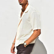 Camicia uomo bianca traforata a maniche corte, stile casual estivo, indossata con pantaloni scuri. Accessori: orologio e bracciali dorati.