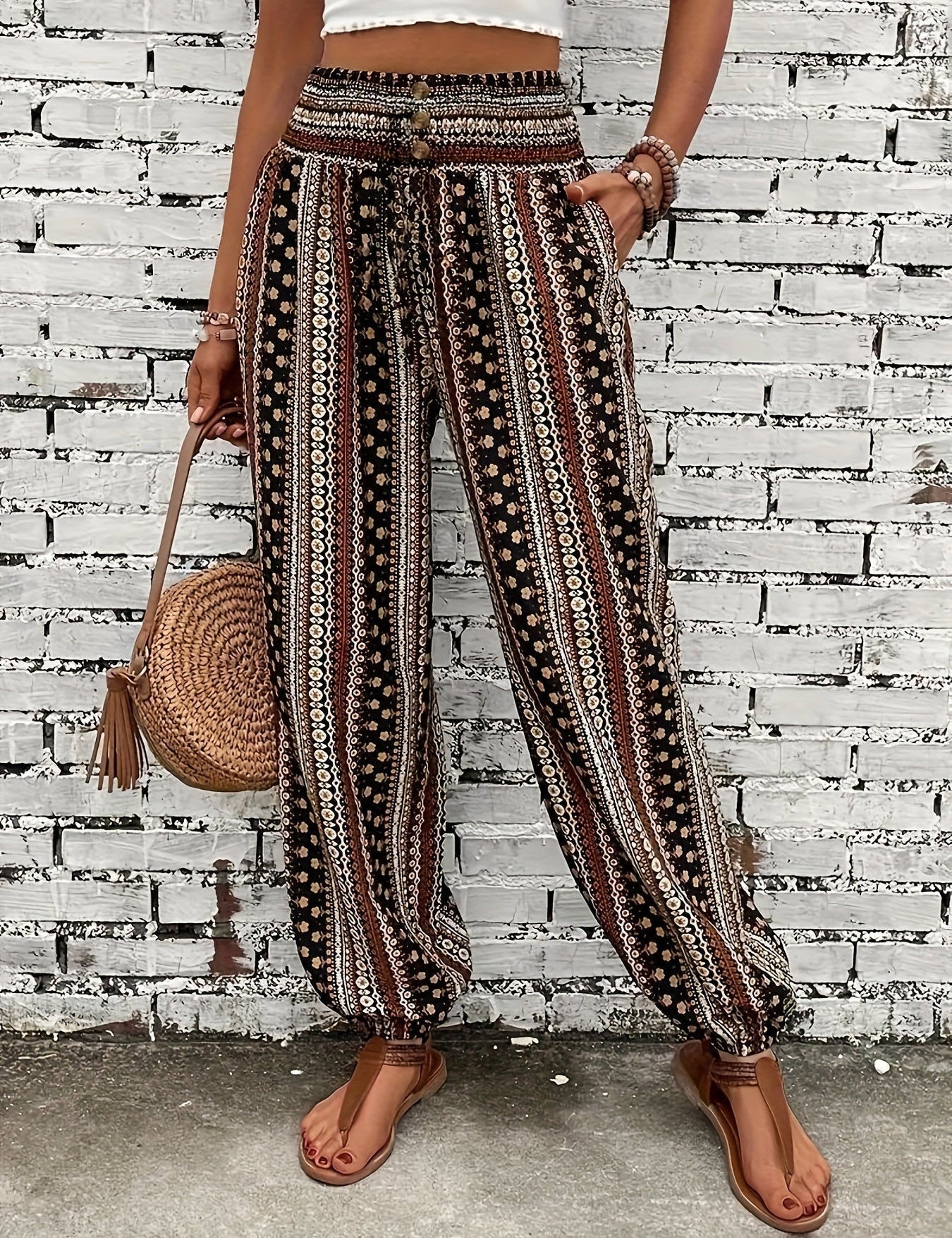 Pantaloni boho a righe etniche, donna, con borsa intrecciata e sandali, moda estiva casual, sfondo muro bianco.
