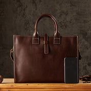Borsa ventiquattrore in pelle marrone con cuciture bianche, manici robusti, accanto a uno smartphone nero su sfondo grigio.