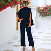 Donna con tuta blu elegante, cappello di paglia e sandali, posa in cortile mediterraneo con fiori rossi. Moda estiva, abbigliamento casual chic.