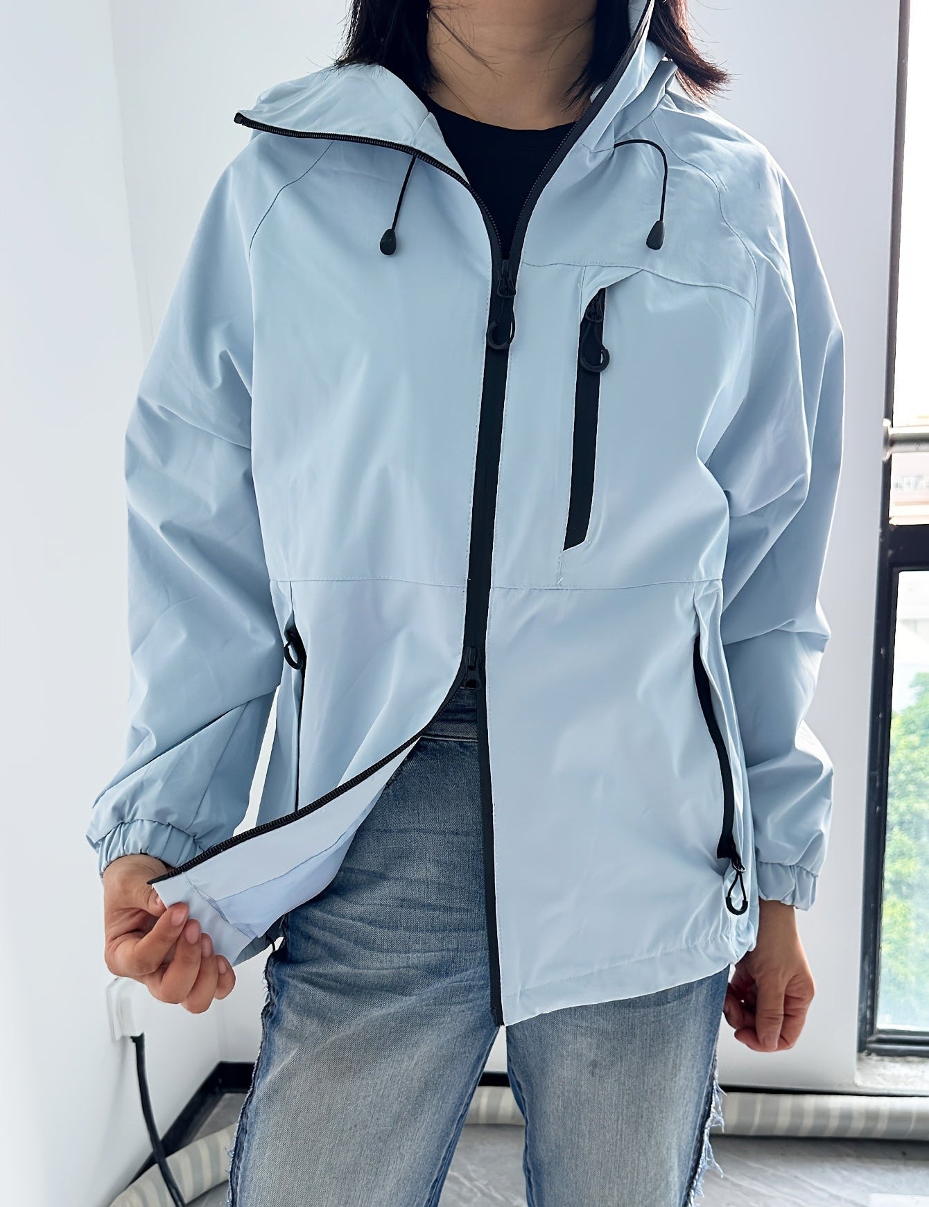 Giacca a vento azzurra da donna con cerniere nere, indossata sopra jeans. Design casual, ideale per attività all'aperto. Tessuto impermeabile.