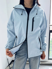 Giacca a vento azzurra da donna con cerniere nere, indossata sopra jeans. Design casual, ideale per attività all'aperto. Tessuto impermeabile.