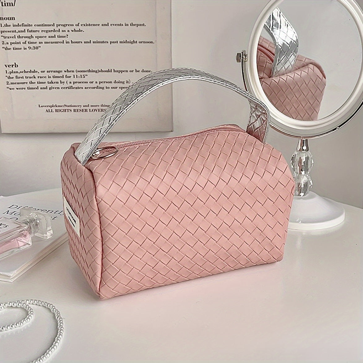 Borsa cosmetica rosa intrecciata su tavolo bianco con specchio rotondo, design elegante e moderno, ideale per organizzare trucchi e accessori.