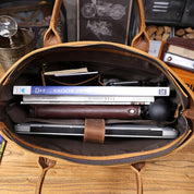 Borsa in pelle marrone aperta con laptop, libri, penna e accessori all'interno, su tavolo in legno. Elegante e pratica per lavoro e studio.