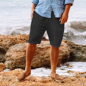 Uomo in pantaloncini neri e camicia azzurra su spiaggia rocciosa, indossa mocassini marroni. Moda estiva casual, abbigliamento maschile.