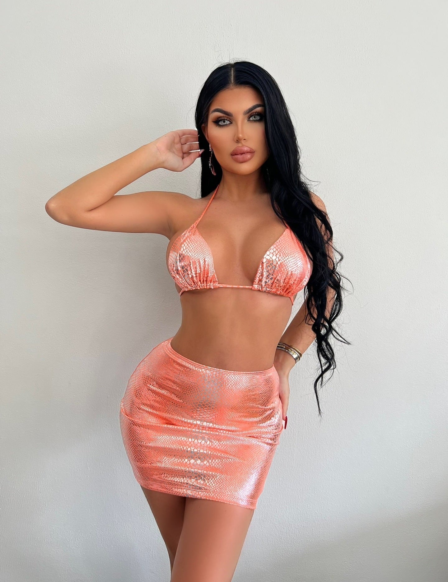 Donna in posa con top bikini e minigonna rosa scintillante, capelli lunghi e neri, sfondo bianco. Moda estiva, abbigliamento glamour.