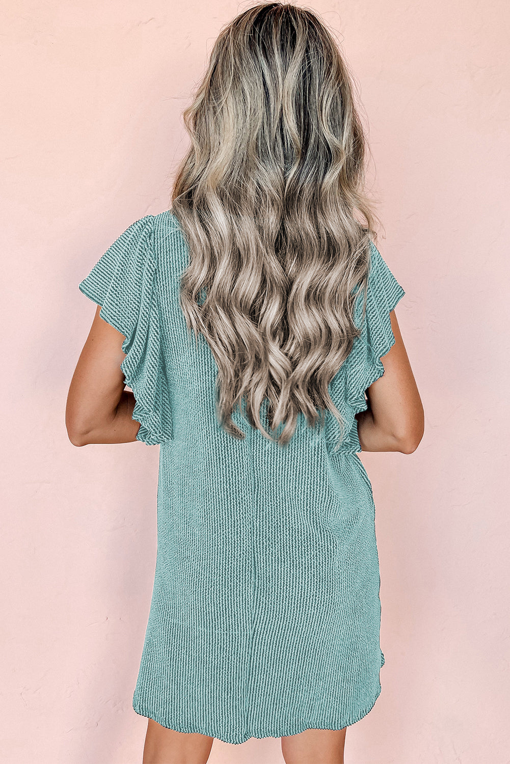 Donna con capelli ondulati biondi indossa un abito verde menta a maniche corte, sfondo rosa. Moda estiva, abbigliamento casual elegante.