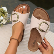 Sandali donna beige con fibbia grande, suola in sughero, indossati su tappeto bianco. Moda estiva, calzature comode, stile casual elegante.