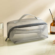 Borsa da toilette trasparente grigia con cerniera, manico superiore, su tavolo bianco. Ideale per viaggi, organizzazione cosmetici e accessori bagno.