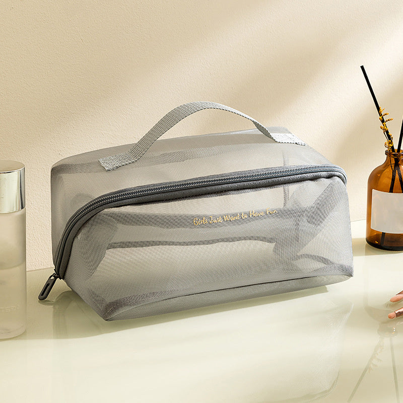 Borsa da toilette trasparente grigia con cerniera, manico superiore, su tavolo bianco. Ideale per viaggi, organizzazione cosmetici e accessori bagno.