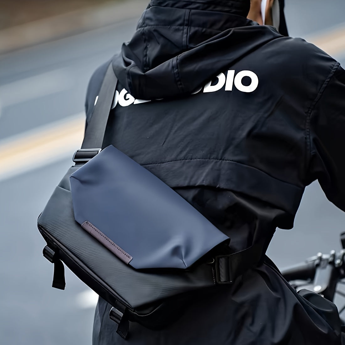 Giacca nera impermeabile con logo bianco, indossata da ciclista. Borsa a tracolla blu scuro, design moderno, ideale per ciclismo urbano.