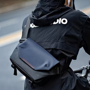 Giacca nera impermeabile con logo bianco, indossata da ciclista. Borsa a tracolla blu scuro, design moderno, ideale per ciclismo urbano.