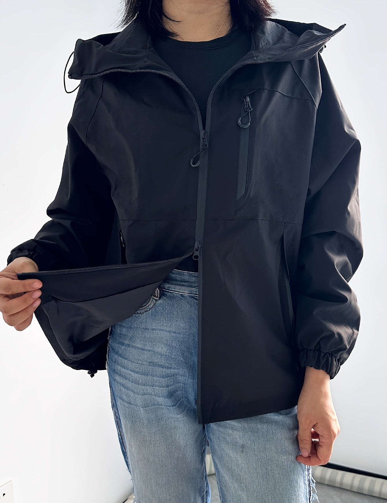 Giacca impermeabile nera da donna con cappuccio, chiusura a zip, tasche multiple, indossata con jeans chiari. Abbigliamento outdoor, moda casual.