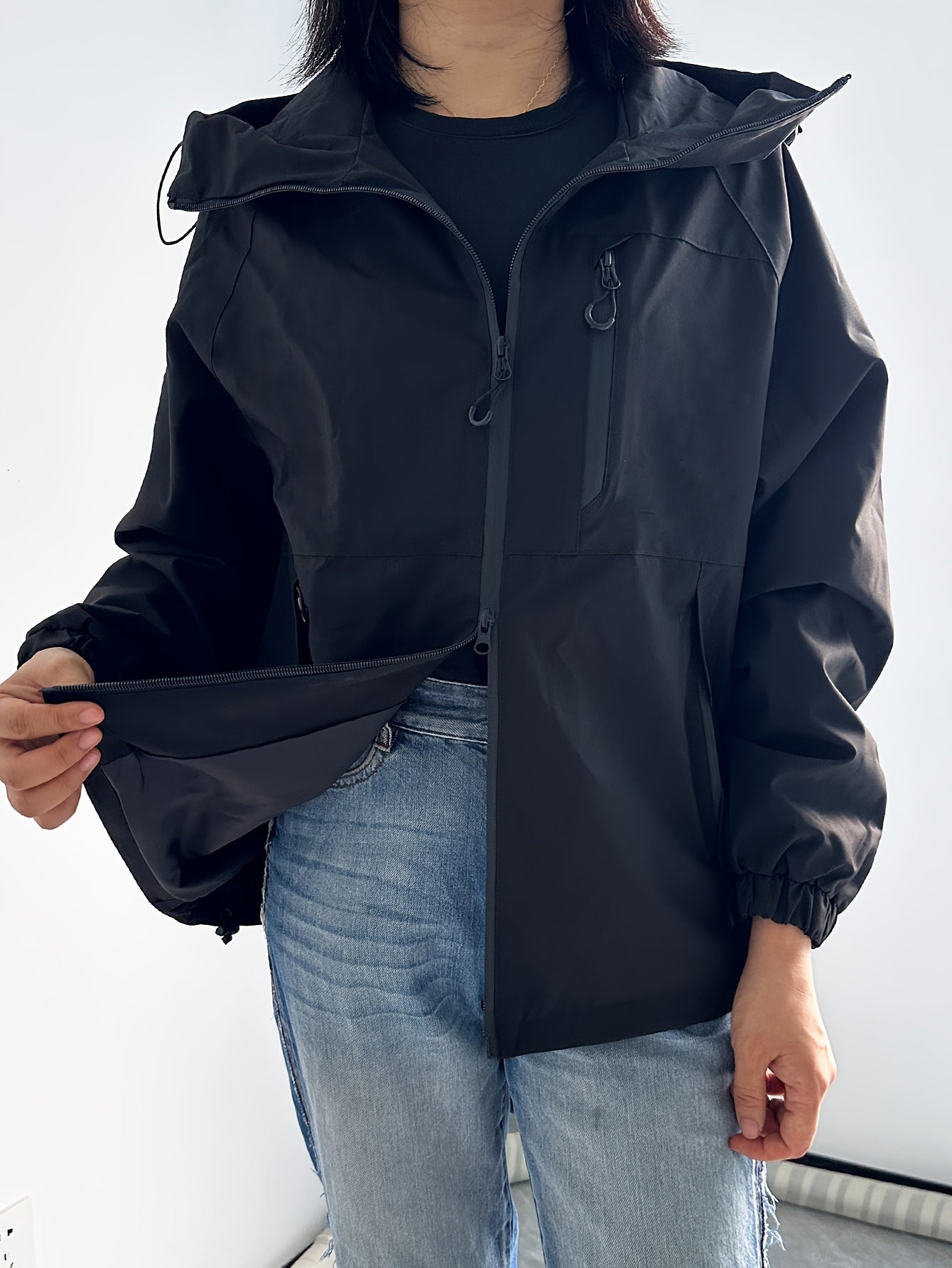 Giacca impermeabile nera da donna con cappuccio, chiusura a zip, tasche multiple, indossata con jeans chiari. Abbigliamento outdoor, moda casual.