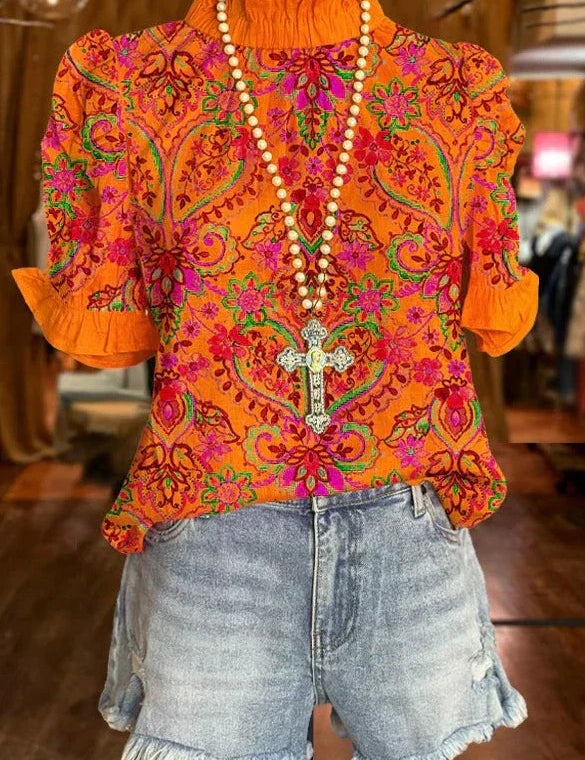 Blusa arancione con motivi floreali rosa e verdi, collana con croce, abbinata a shorts di jeans sfrangiati. Moda estiva, abbigliamento casual donna.
