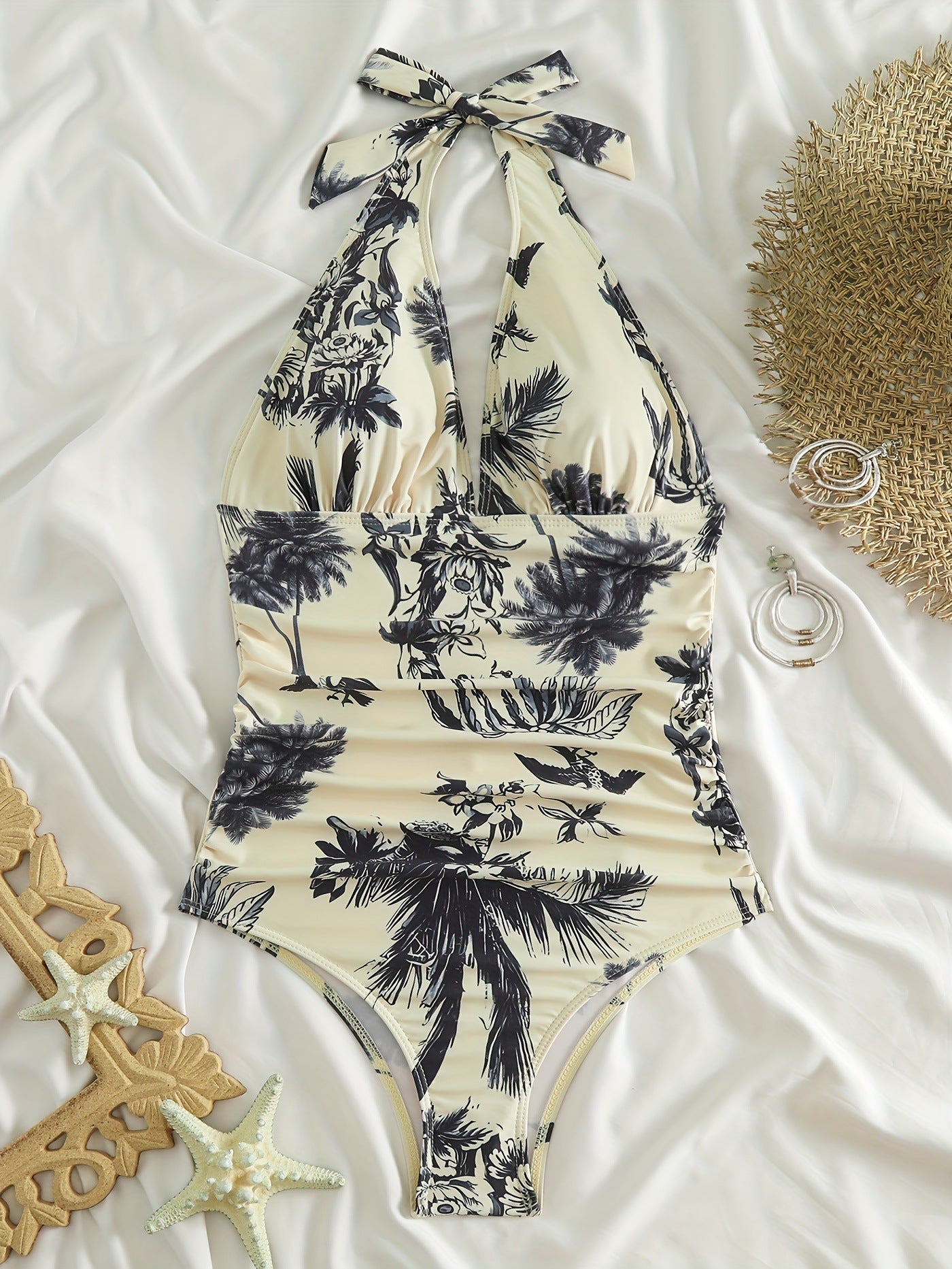 Costume intero donna con stampa tropicale bianco e nero, su sfondo di tessuto bianco. Accessori estivi: cappello di paglia, orecchini e stelle marine.