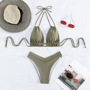 Bikini verde oliva a due pezzi su sfondo bianco, con cappello a righe, occhiali da sole rossi e rivista. Moda mare estiva, accessori spiaggia.