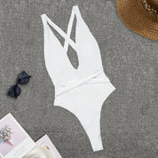 Costume intero bianco da donna con scollo a V incrociato, su sfondo grigio. Accessori: occhiali da sole, cappello di paglia, rivista.