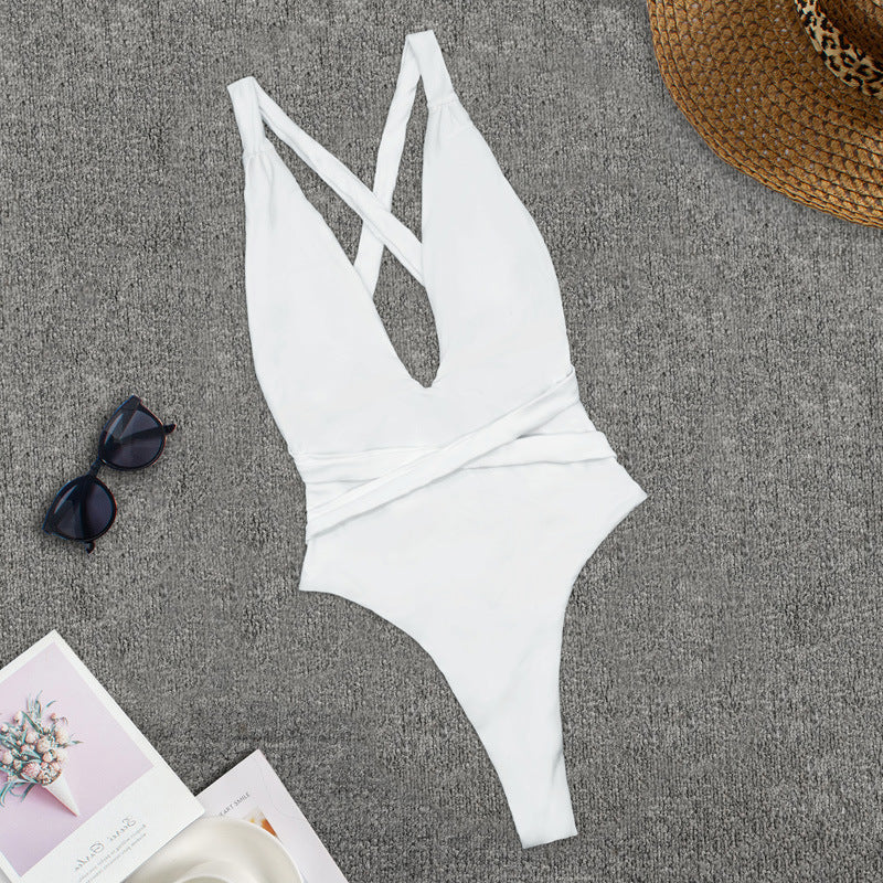 Costume intero bianco da donna con scollo a V incrociato, su sfondo grigio. Accessori: occhiali da sole, cappello di paglia, rivista.