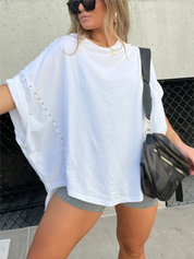 Donna con maglietta bianca oversize con borchie, pantaloncini grigi, borsa a tracolla nera, occhiali da sole. Moda casual estiva urbana.