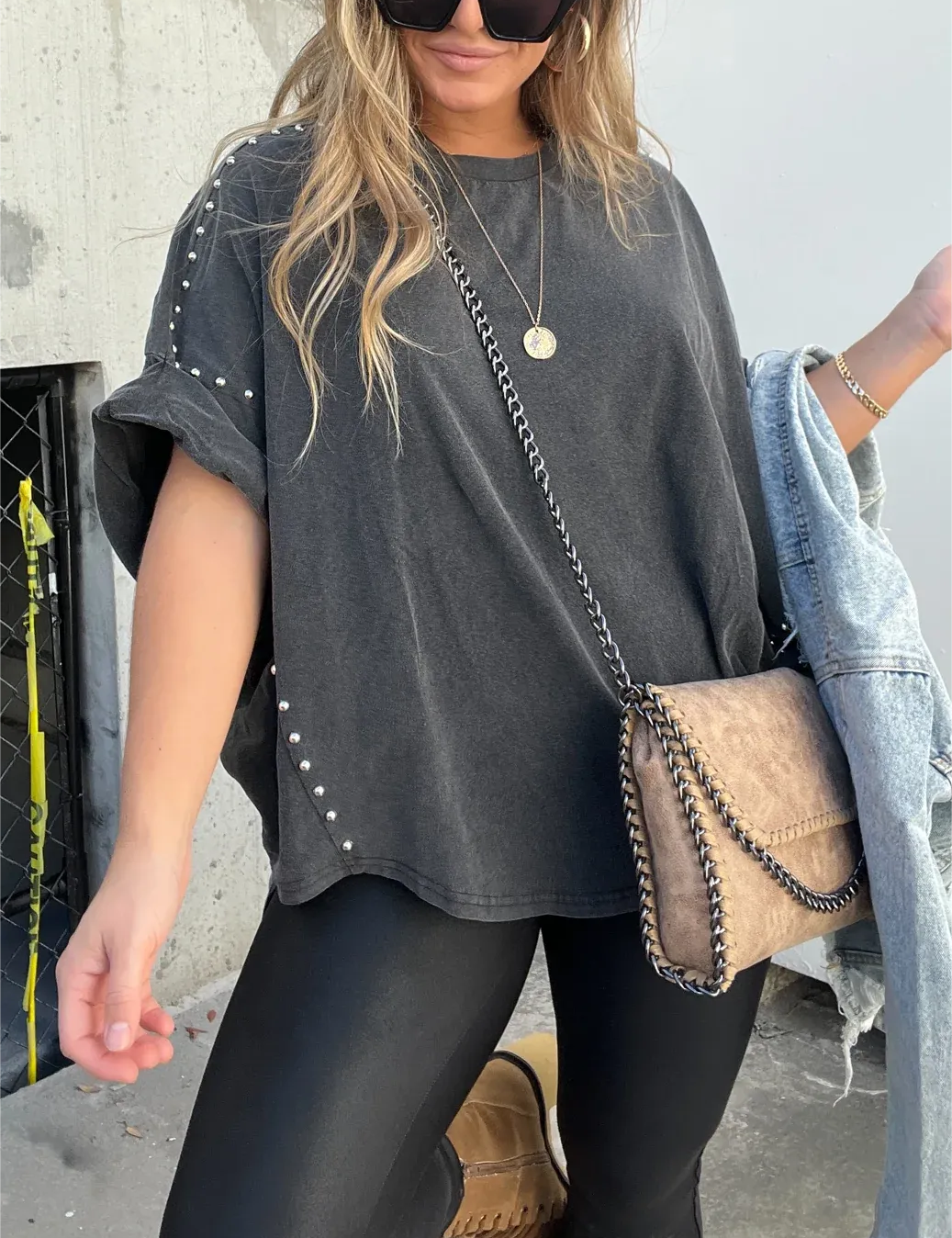 Donna con maglietta oversize nera con borchie, leggings neri, borsa a tracolla beige, occhiali da sole e giacca di jeans. Moda casual urbana.