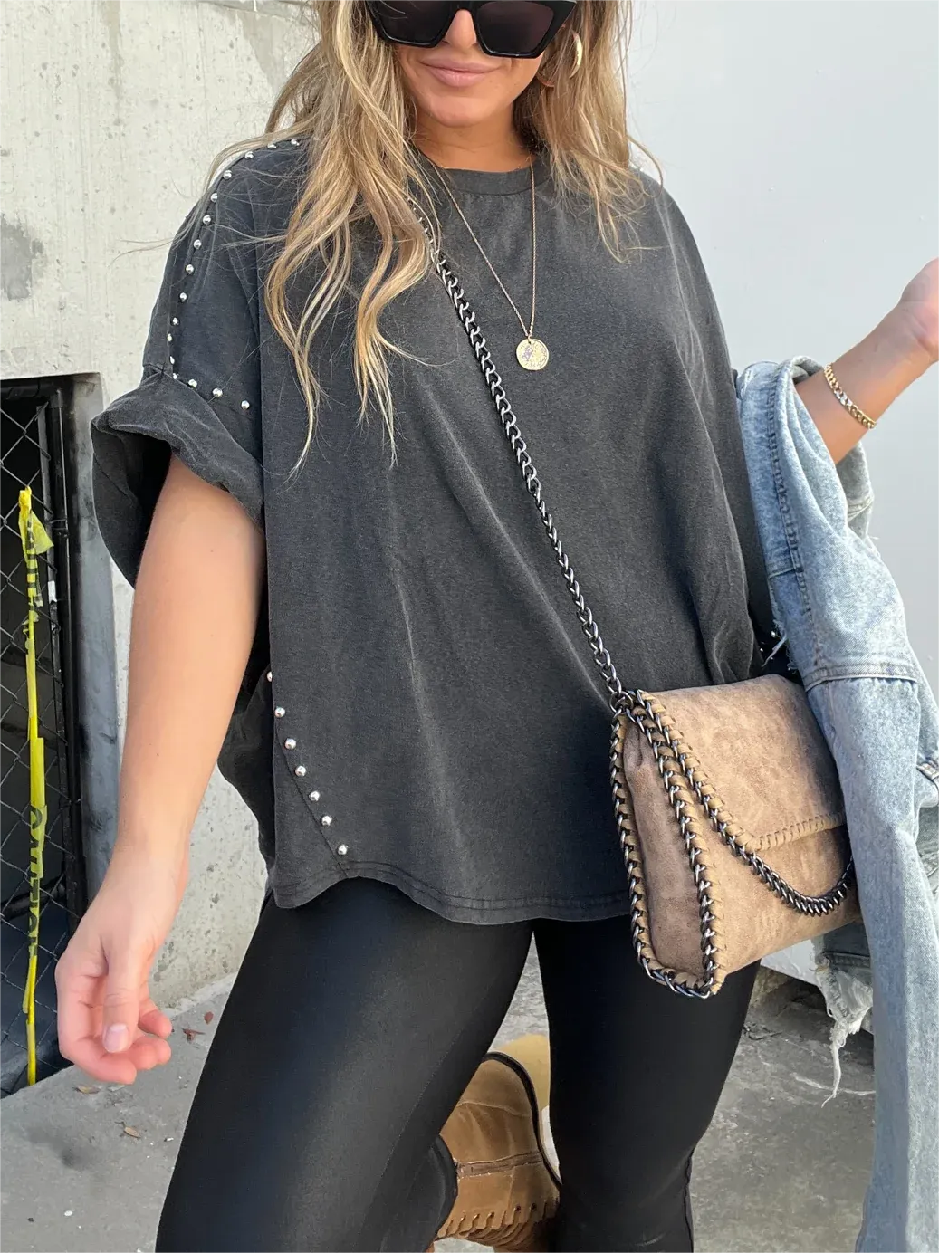 Donna con maglietta oversize nera con borchie, leggings neri, borsa a tracolla beige, occhiali da sole e giacca di jeans. Moda casual urbana.