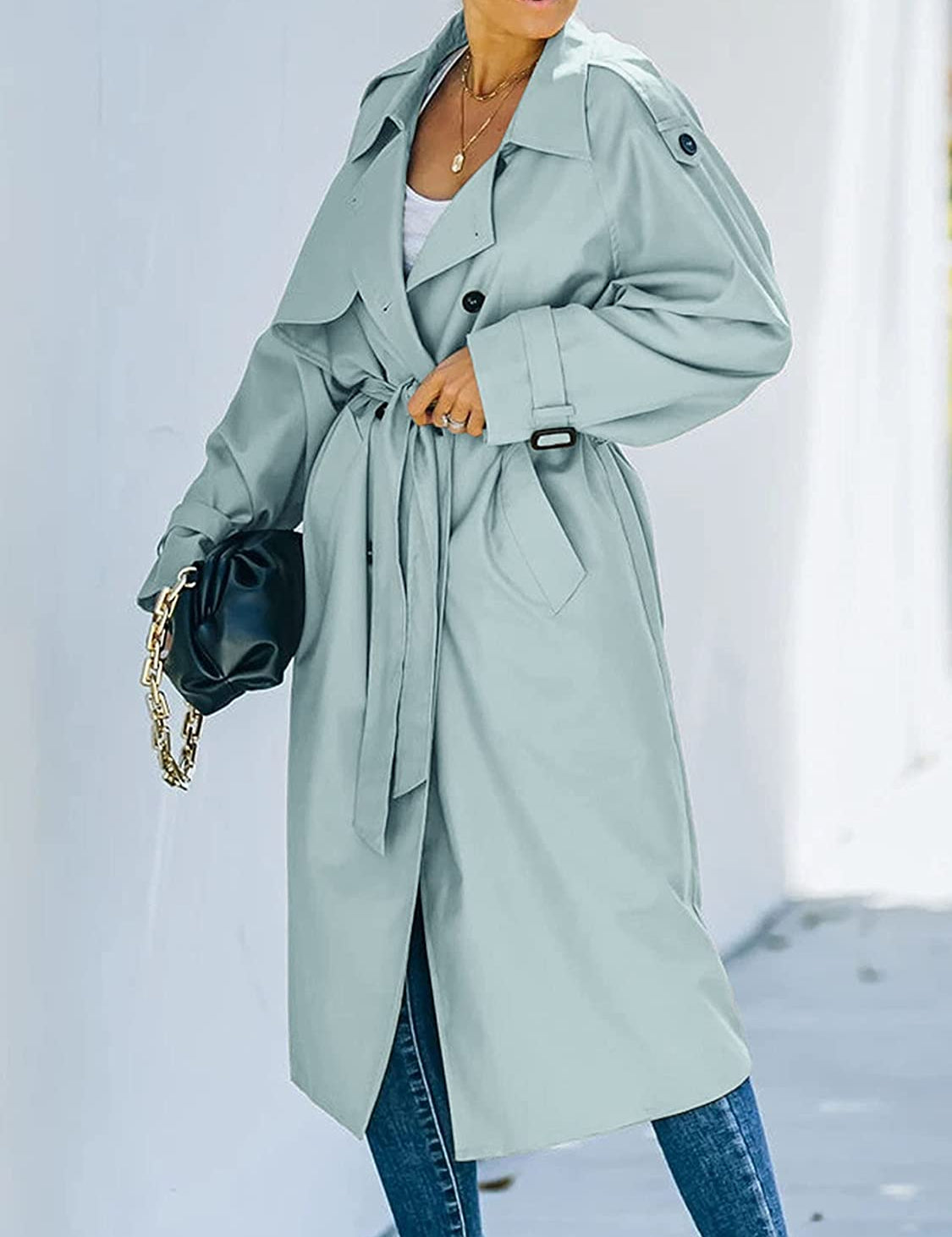 Cappotto trench donna elegante azzurro, cintura in vita, tasche laterali, abbinato a jeans e borsa nera. Moda autunnale chic.