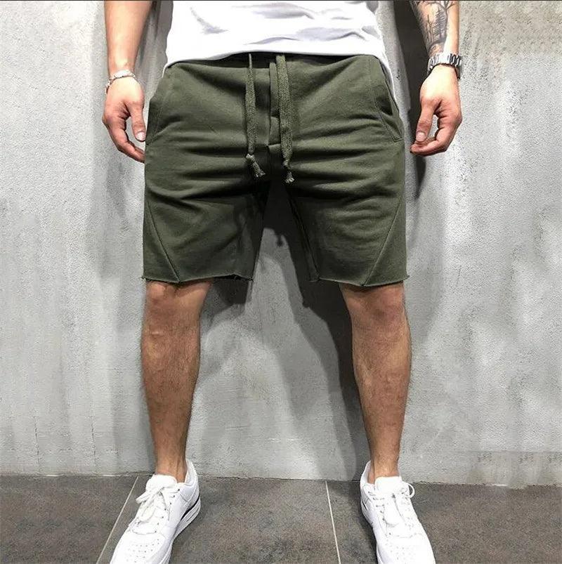 Uomo in pantaloncini verdi casual, scarpe da ginnastica bianche, posa contro un muro grigio. Moda estiva maschile, abbigliamento sportivo trendy.