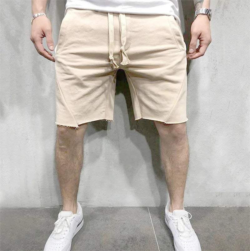 Pantaloncini beige da uomo in cotone, con orlo sfrangiato e tasche laterali, indossati con scarpe da ginnastica bianche. Moda casual estiva.