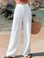 Pantaloni bianchi larghi in lino su spiaggia, moda estiva donna, abbinati a borsa di paglia. Look casual chic per vacanze al mare.