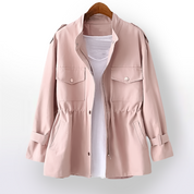 Giacca rosa chiaro da donna con tasche frontali e cintura, appesa su gruccia. Design casual, ideale per moda primaverile.