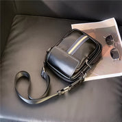 Borsa a tracolla in pelle nera su divano, con striscia decorativa blu e beige, accanto a occhiali da sole e rivista. Accessorio moda elegante.