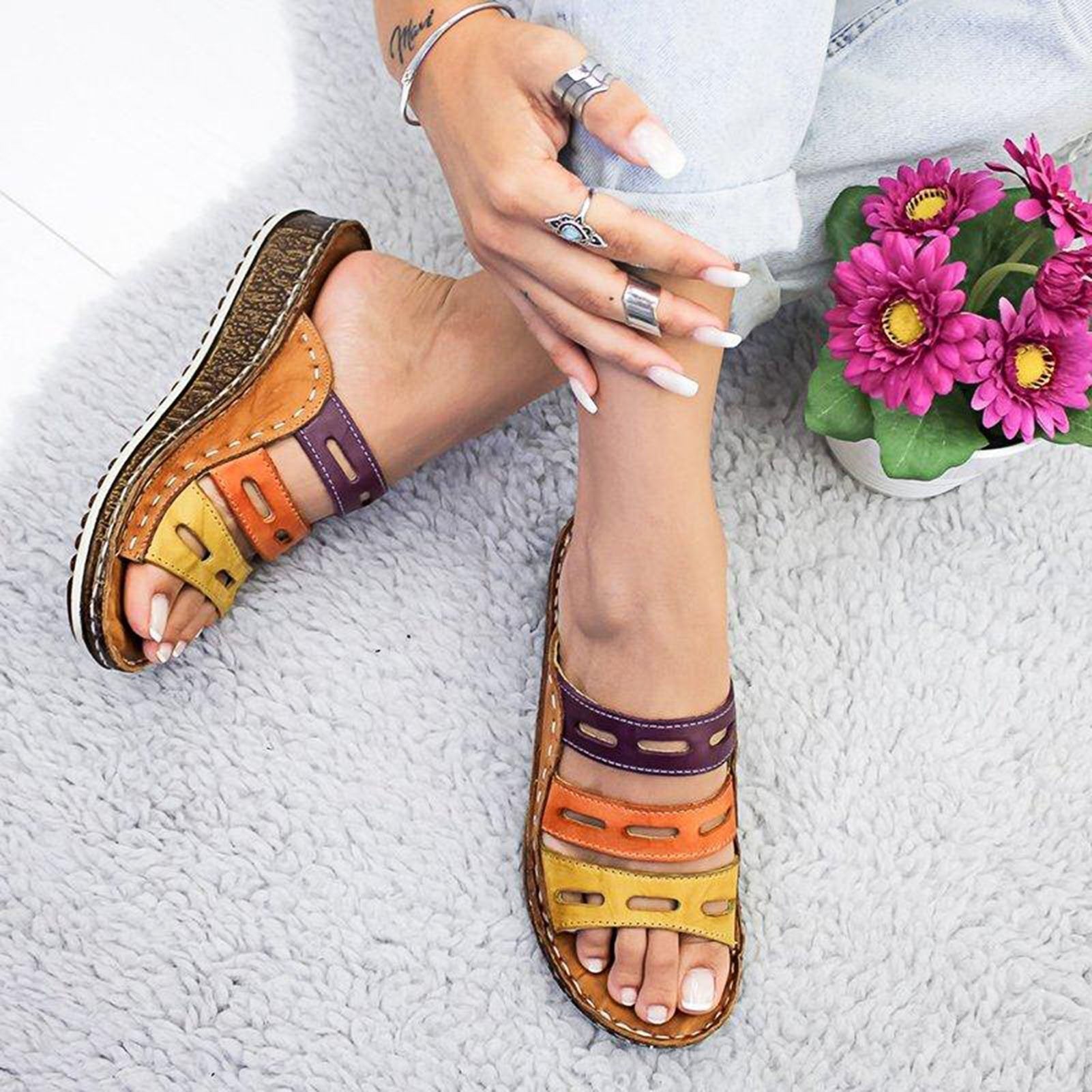 Donna con sandali multicolore in pelle su tappeto grigio, fiori rosa accanto. Scarpe estive casual, moda femminile, comfort e stile.