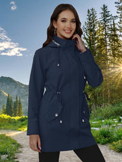 Giacca impermeabile blu donna, cappuccio regolabile, tasche con bottoni, perfetta per escursioni in montagna. Sfondo natura, sole tra alberi.
