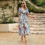 Donna con abito floreale lungo, occhiali da sole e sandali bianchi, cammina su un sentiero di pietra. Moda estiva elegante, stile casual chic.