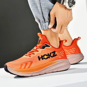 Scarpe da corsa arancioni HOKZ, design sportivo, suola ammortizzata, logo nero, indossate con pantaloni grigi. Ideali per allenamento e jogging.