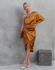 Donna in abito monospalla color bronzo, elegante e drappeggiato, con tacchi bianchi e pochette marrone, posa su sfondo grigio moderno.