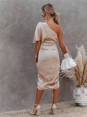 Donna in abito elegante monospalla beige, sandali con tacco, borsa bianca, sfondo neutro. Moda femminile, abbigliamento chic, stile sofisticato.