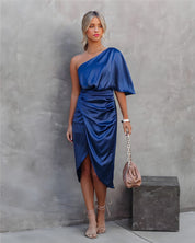 Donna in abito blu monospalla elegante, con drappeggio e sandali beige, posa contro un muro grigio. Borsa a catena rosa tenue. Moda femminile chic.