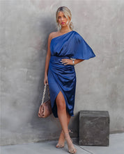 Donna in abito blu monospalla elegante, posa contro un muro grigio. Indossa sandali beige e tiene una borsa marrone. Moda femminile, abbigliamento elegante.