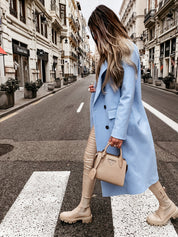 Donna con cappotto azzurro e borsa beige cammina su strada cittadina. Moda autunnale, stivali beige, look urbano elegante.