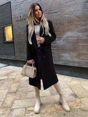 Donna con cappotto nero e stivali beige, borsa Prada, posa su marciapiede urbano. Moda autunnale, stile elegante, capelli lunghi biondi.