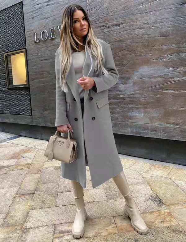 Donna con cappotto grigio elegante, borsa beige e stivali abbinati, posa davanti a un muro moderno. Moda autunnale, stile urbano chic.